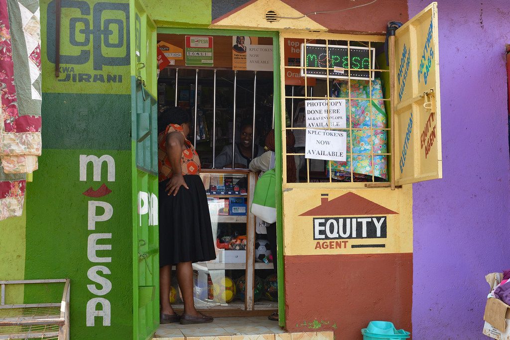 A brightly painted M-Pesa agent storefront in Nairobi with a customer at the counter